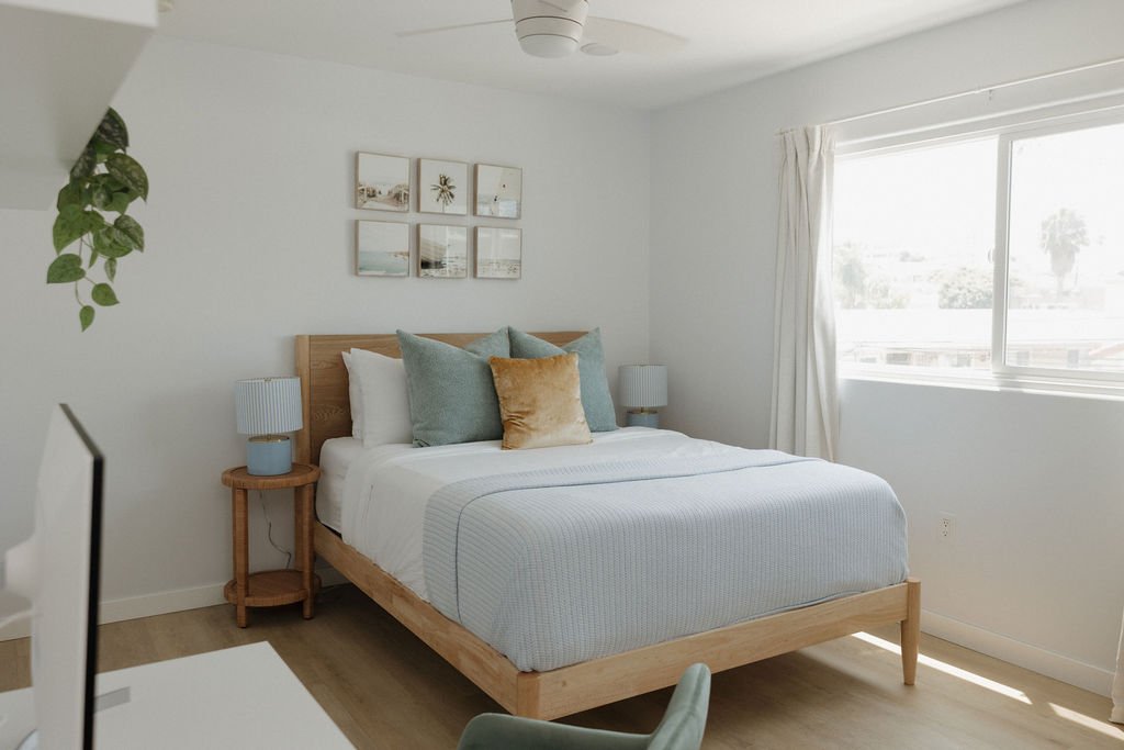 A bedroom with a wooden bed frame, white bedding, and decorative pillows, two nightstands with lamps, a window with white curtains, and framed wall art above the bed.