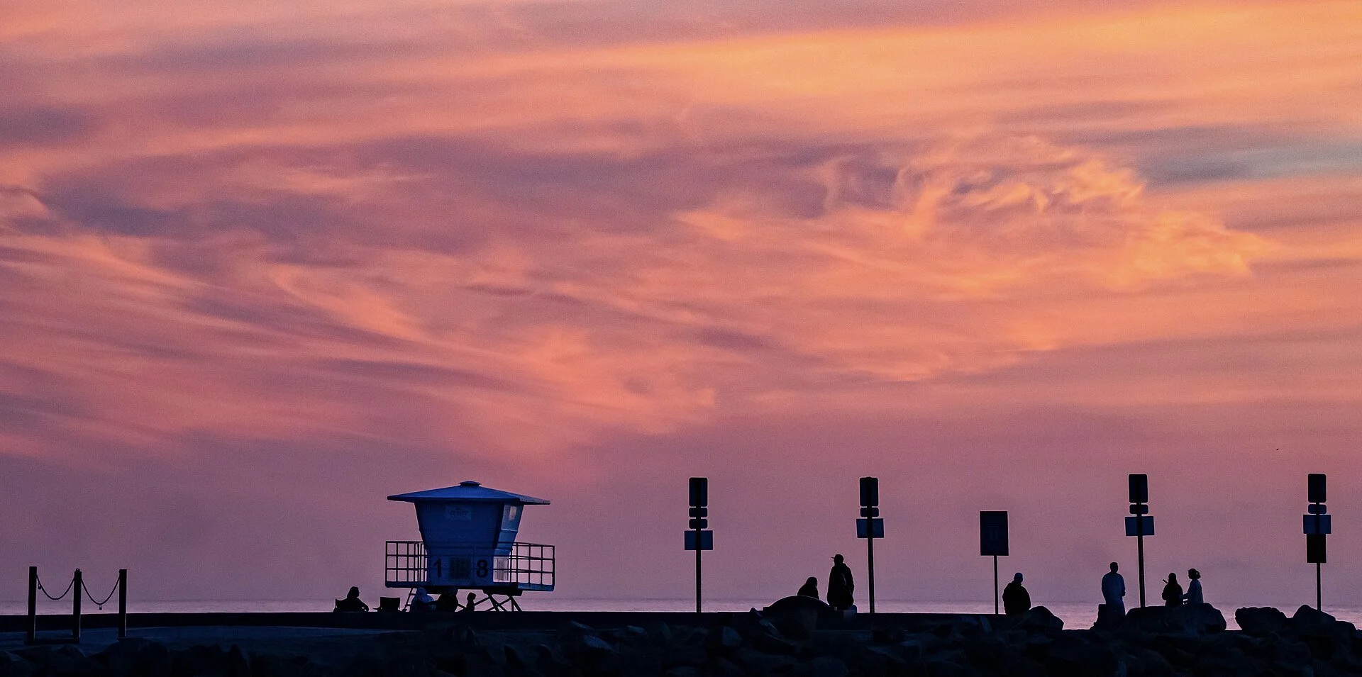 Lifeguard Tower 18 in Oceanside in silhouette against a pink and orange sunset sky, with people watching from the jetty