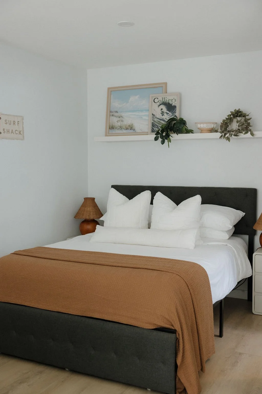 A neatly made bed with white bedding and multiple pillows, topped with a brown blanket, in a minimalist bedroom with white walls. There are two wicker lamps, a shelf above the bed with framed art, a plant, and decorative items.