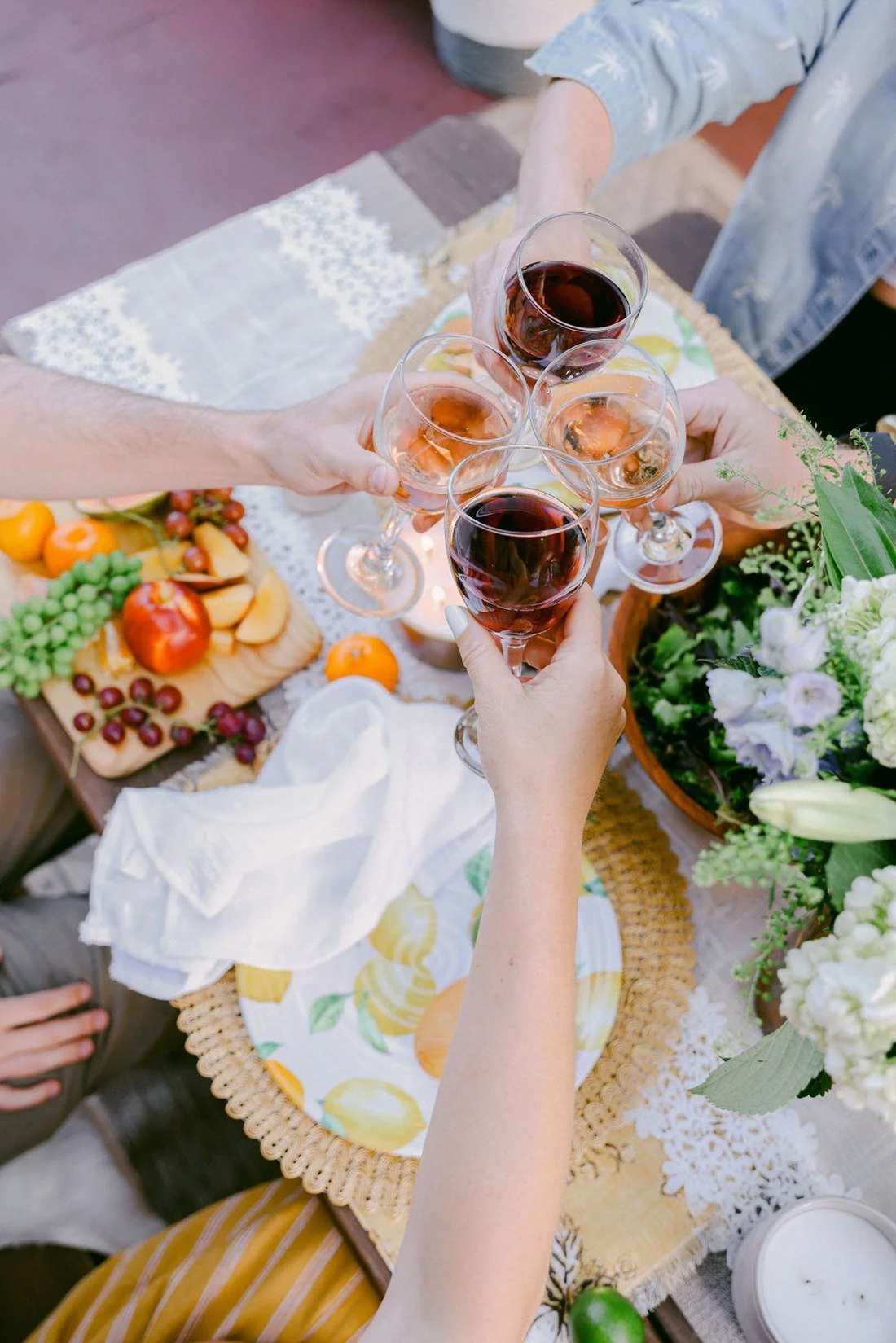 Four glasses of wine raised over a table of fruit, cheese and flowers at sunset