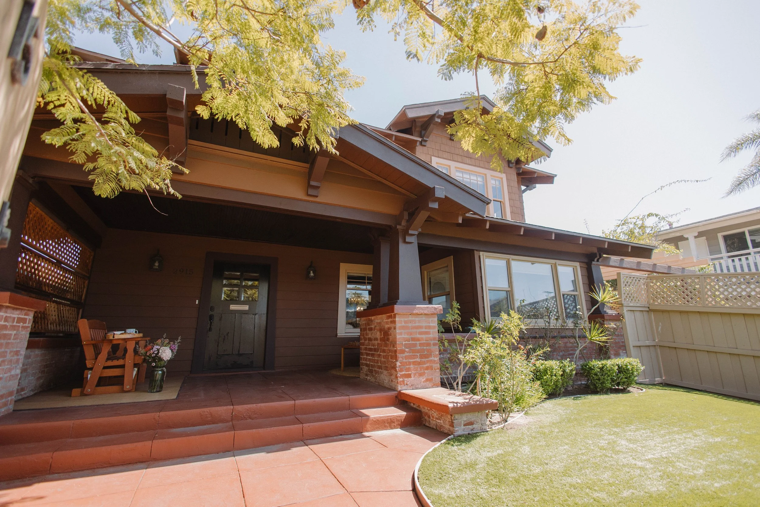 The front porch of Palm Parkside, a 1911 Craftsman near Balboa Park, with brick pillars, a lawn and a jacaranda tree overhead