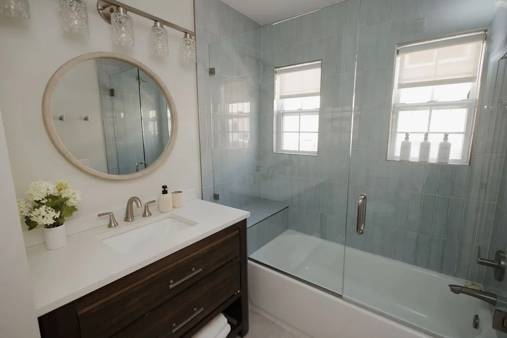 A bathroom in The Villa with a wood vanity, a round mirror and a glass tub and shower