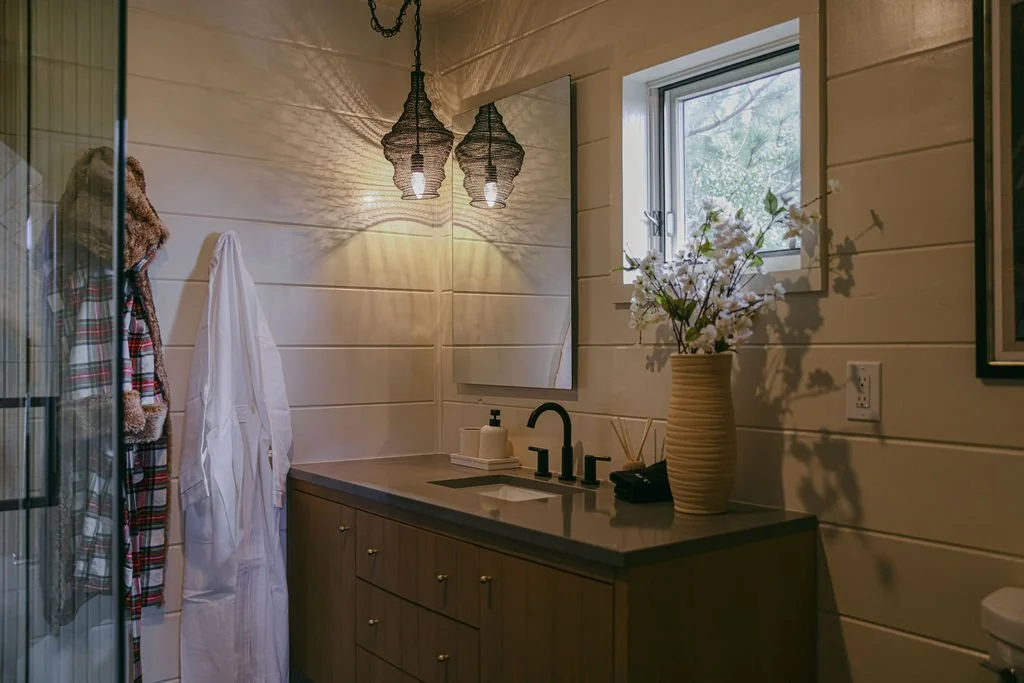 Bathroom with a wooden vanity, large mirror, white flowers in a vase, small window, and hanging light fixtures.