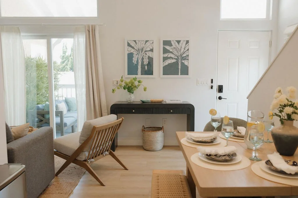 Living and dining area with sliding glass door to patio, light wood flooring, beige and gray furniture, wall art with banana leaf prints, dining table set with plates, glasses, and napkins, and a black console table against the wall with a vase of fl