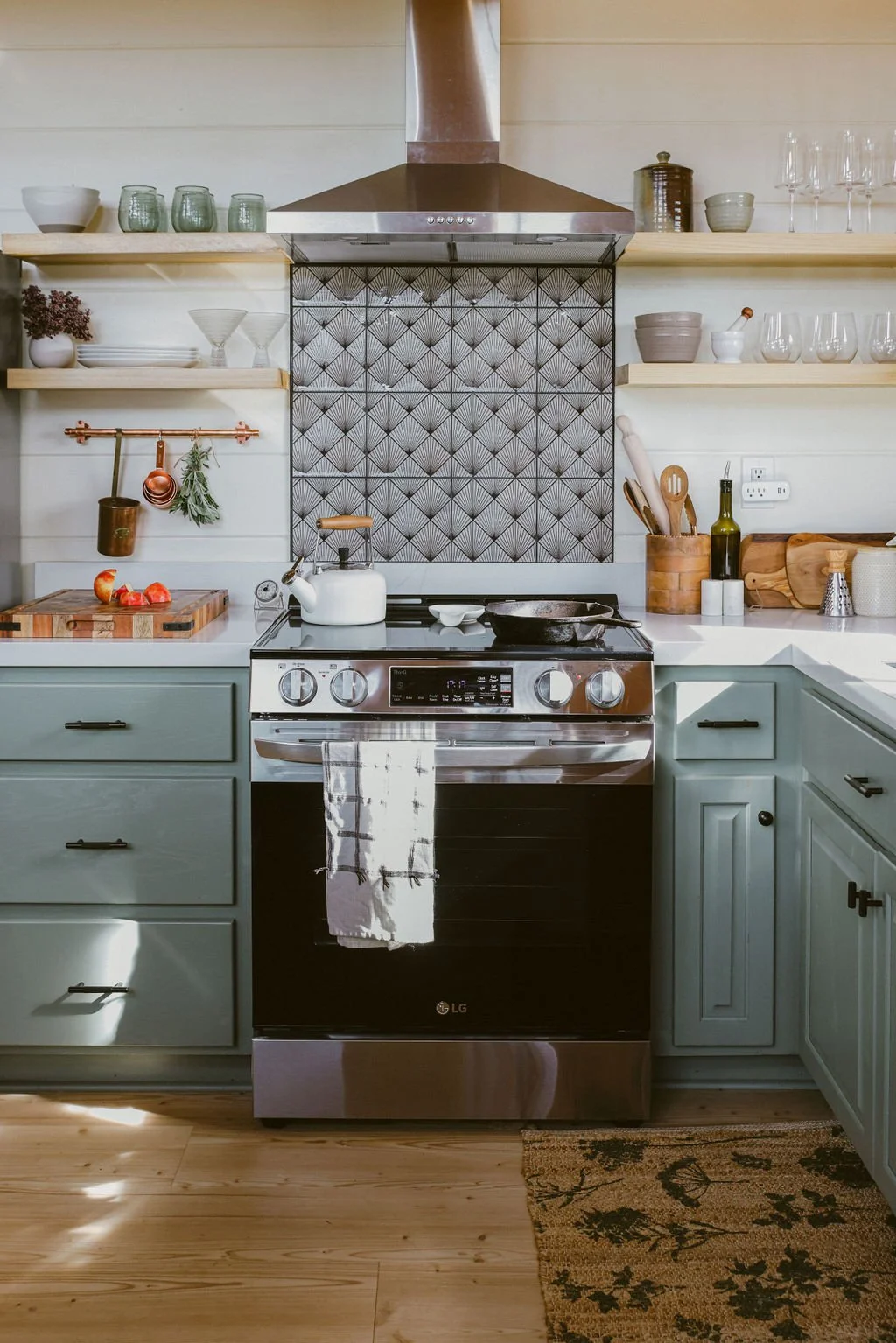 Kitchen with sage green cabinets, stainless steel stove with a white kettle, open shelving with glassware, bowls, cutting boards, cooking utensils, and decorative items, a decorative black and white backsplash, and a wooden floor with a patterned rug.