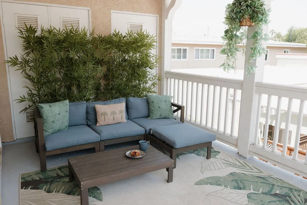 Outdoor porch with a blue cushioned sectional sofa, pillows, a coffee table with a cup and snack, and hanging plants with a white railing and neighboring houses in the background.