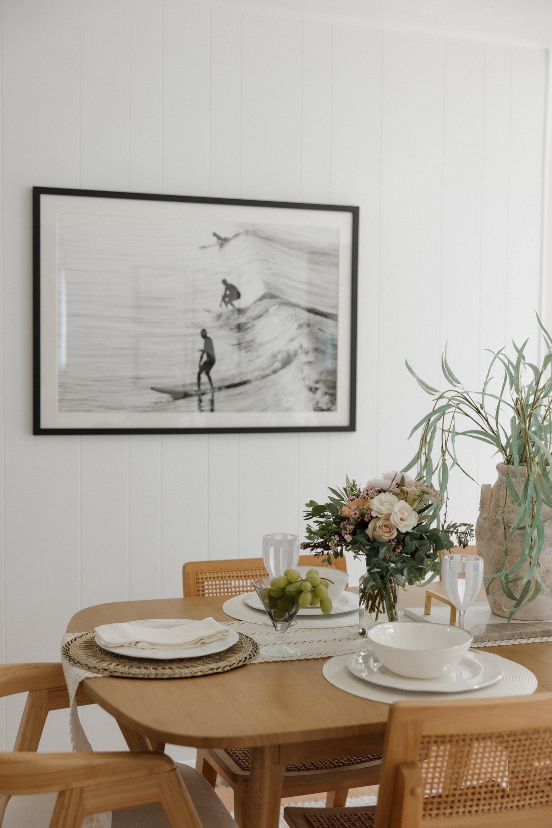 A dining table set with linen, woven placemats, grapes and a vase of roses, beneath a framed black and white surf print