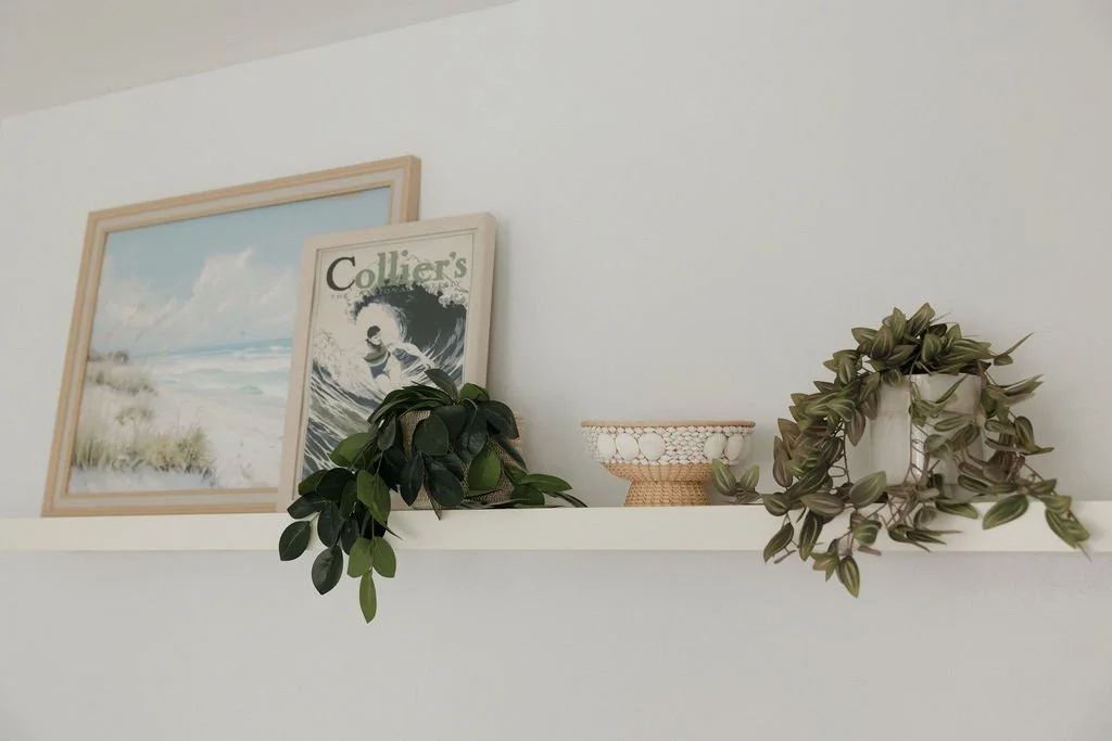 Decorative shelf on a white wall holding a framed picture, a magazine, a potted plant, and a woven bowl.