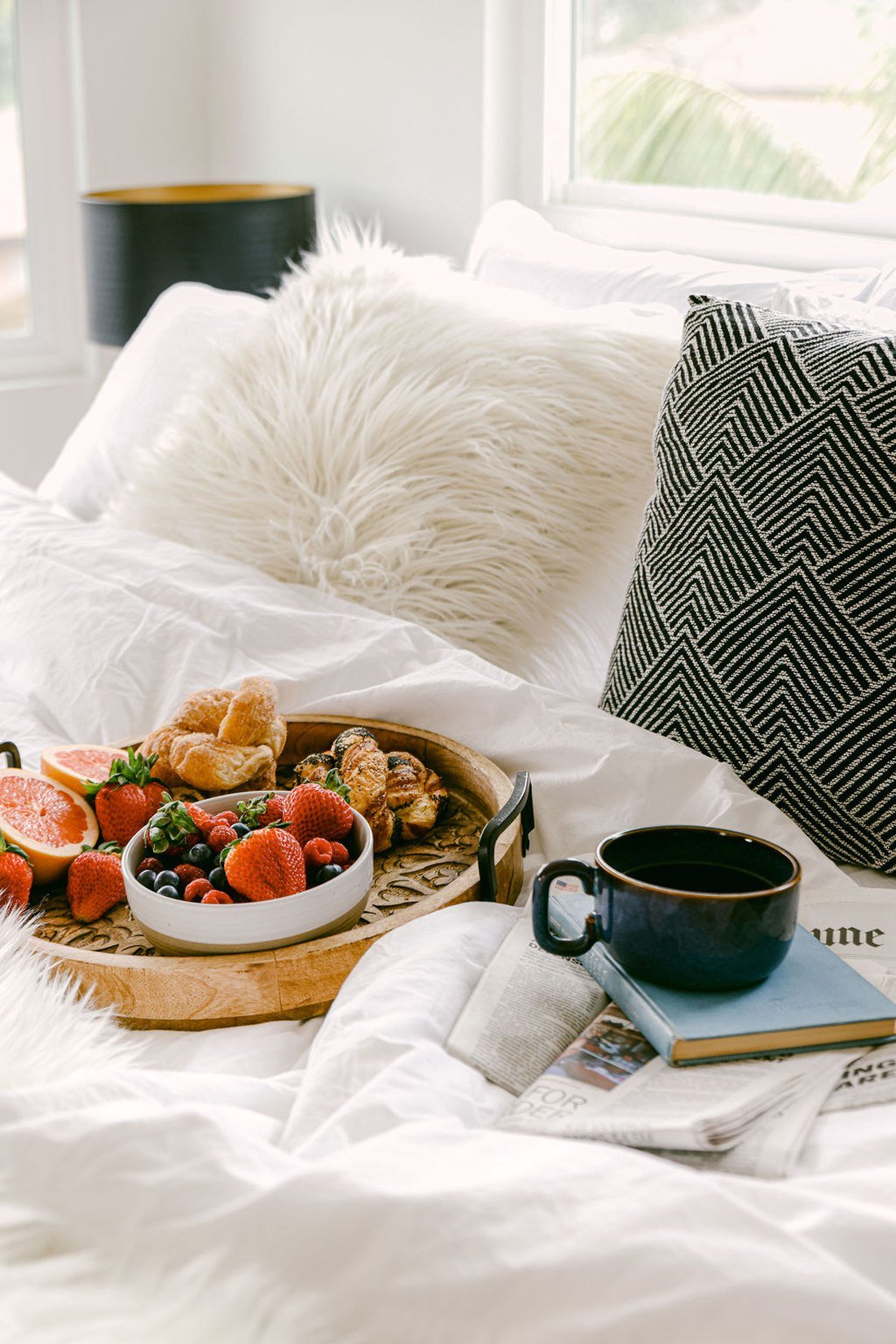 A breakfast tray on white linen in bed, with berries, pastries, a mug of coffee and the morning paper