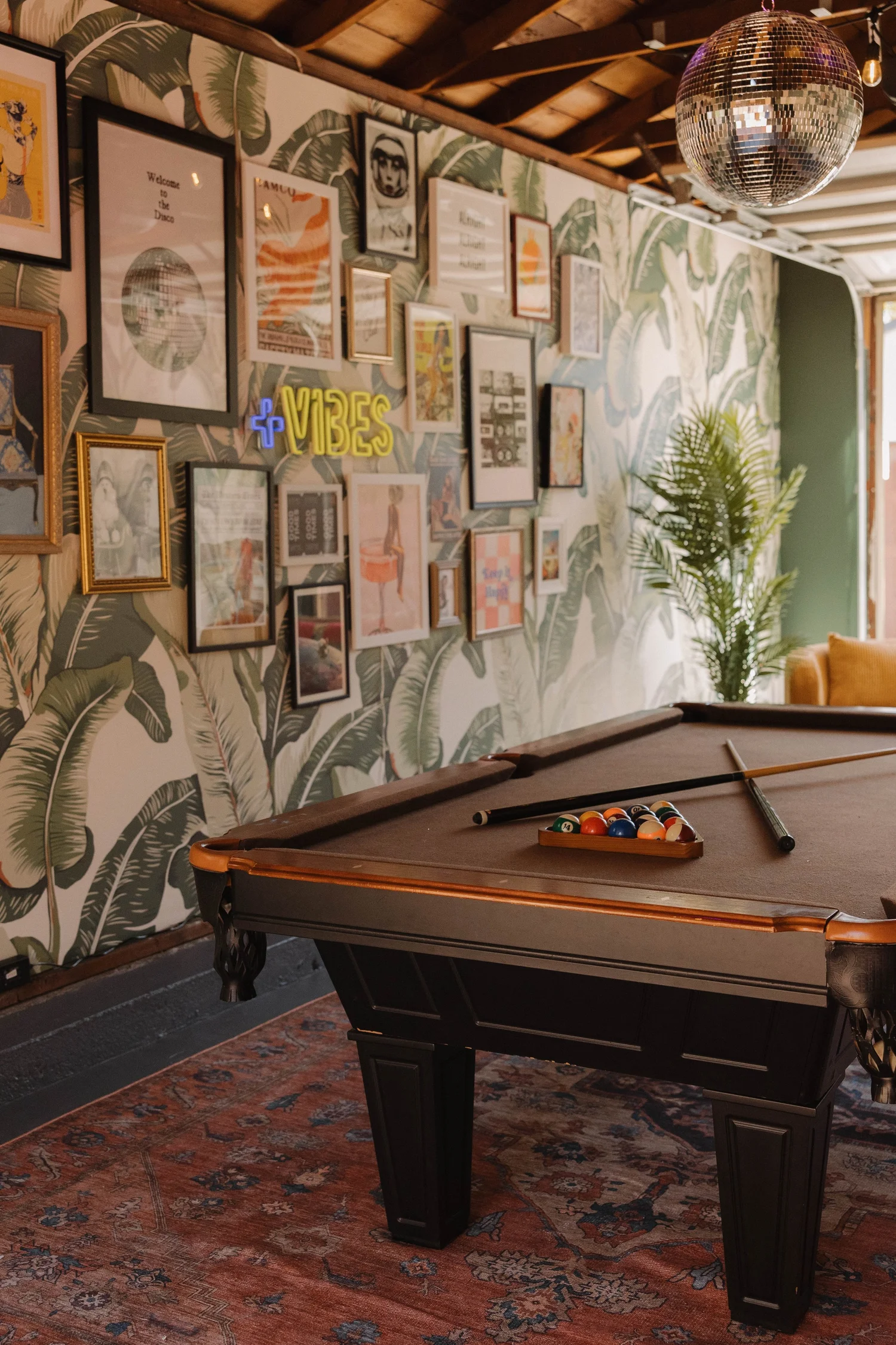 The pool table under a disco ball in the garage game room at Palm Parkside, with palm leaf wallpaper and a wall of framed art