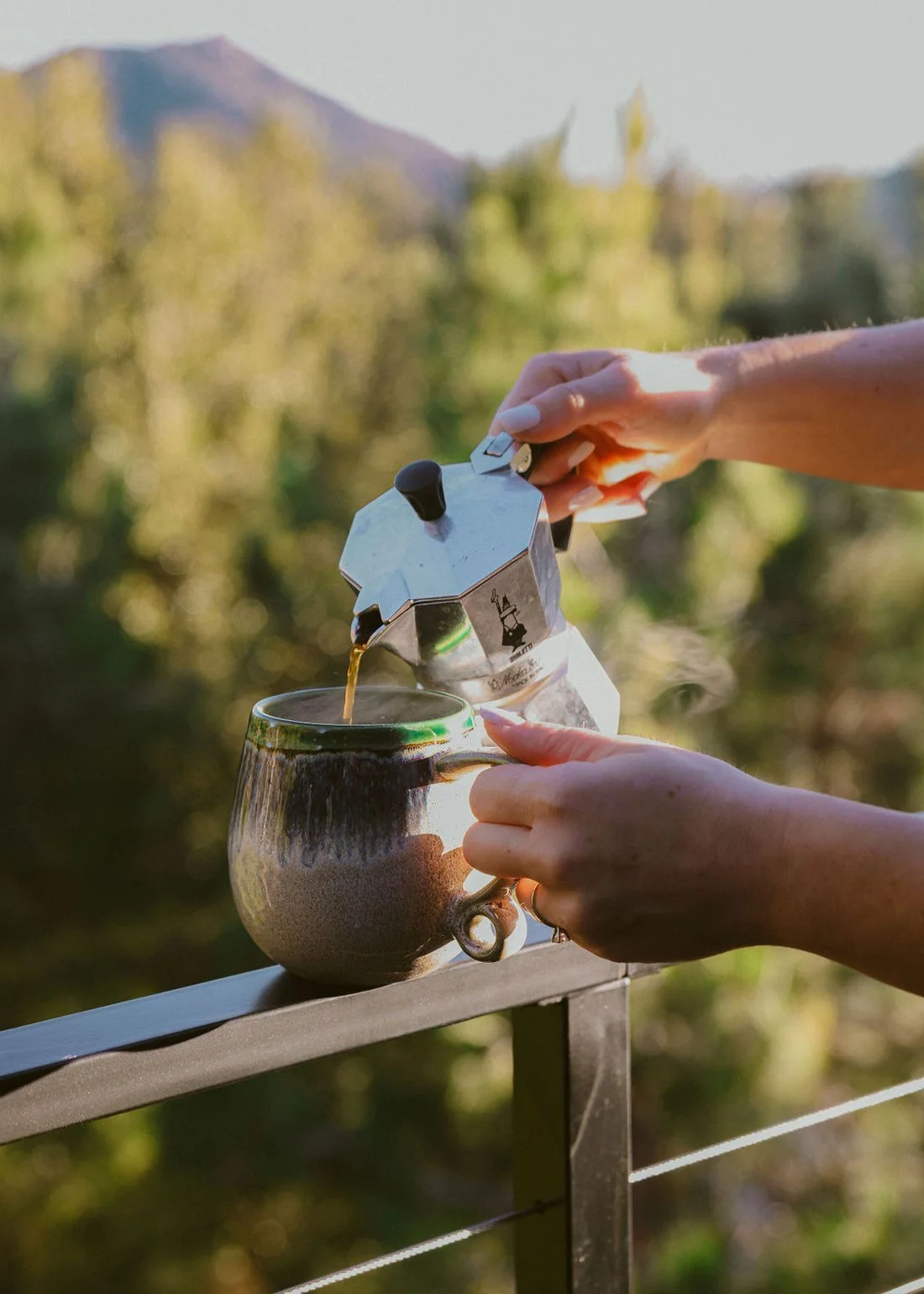 Pouring coffee from a moka pot into a stoneware mug on the deck rail, with the Julian mountains behind