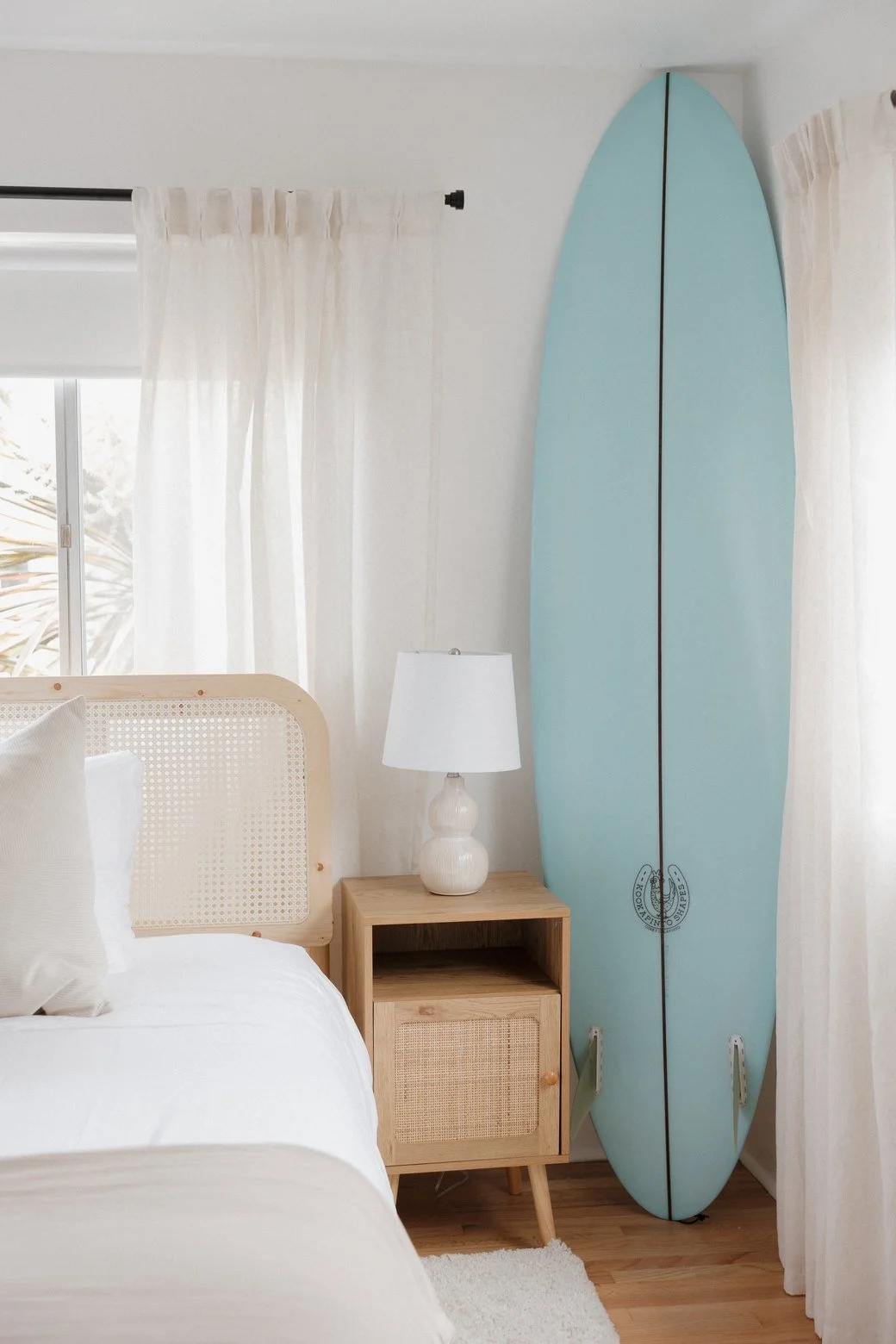 A bright coastal bedroom with a cane headboard, linen curtains and a mint surfboard leaning against the wall