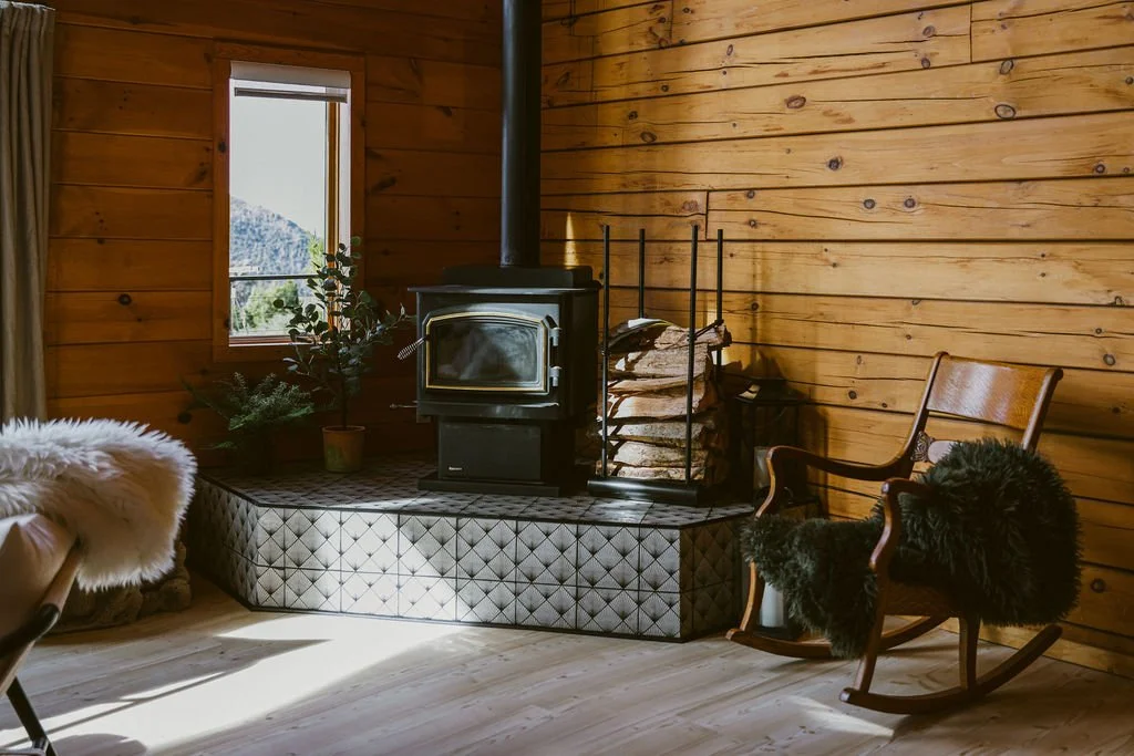 Wood stove on a patterned tile hearth with a rocking chair and sheepskin throws at Dark Moon Chalet in Julian, designed by Salt and Sky Lodging Co.