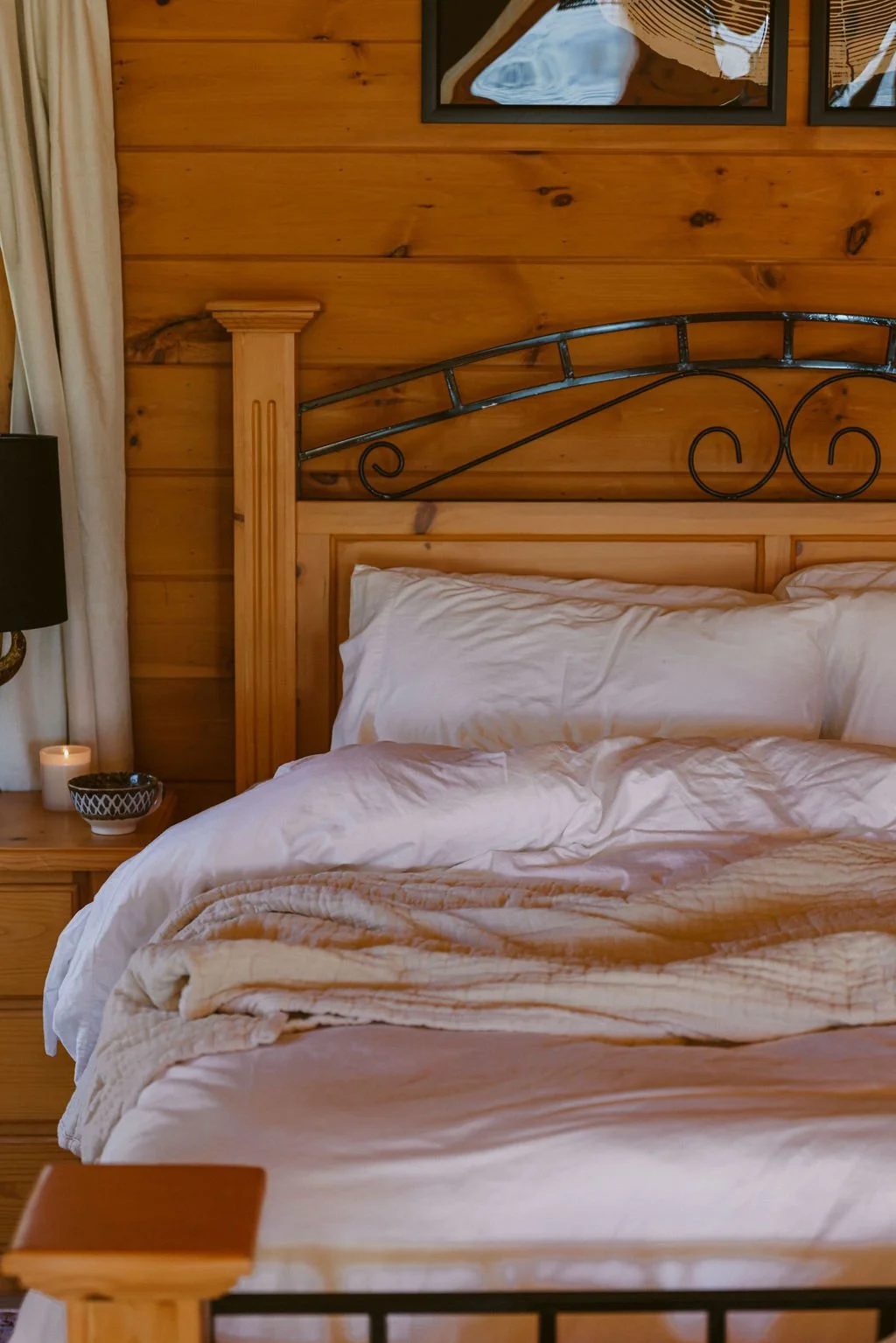 A bed with white bedding in a rustic wood-paneled room, next to a nightstand with a candle and a bowl, and framed artwork above.