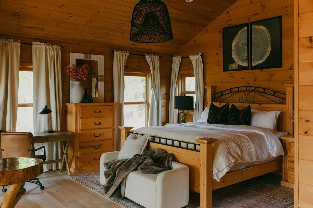 Cozy bedroom with wooden walls and ceiling, large bed with white linens and black pillows, dresser with flowers, desk with lamp, window with cream curtains, small sofa with blanket, and framed wall art.