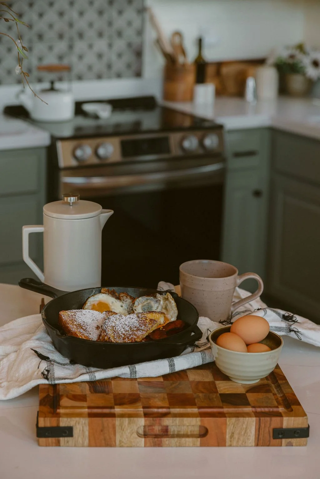 Fried eggs and toast with powdered sugar on a skillet, served on a wooden cutting board with a bowl of eggs and a mug, on a kitchen counter.