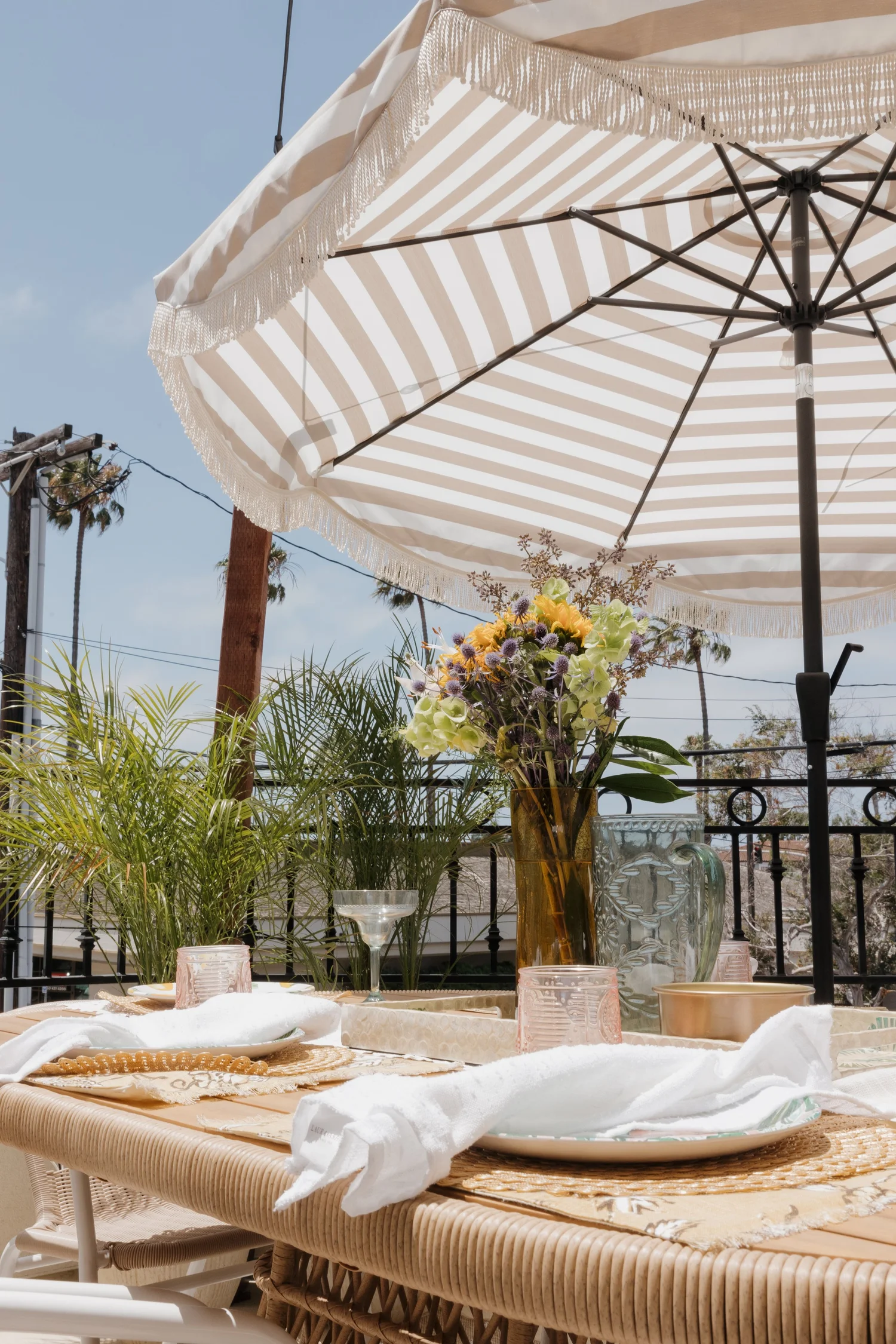 Outdoor dining table under a striped umbrella with flowers and palms at Coastal Palm in Pacific Beach, designed by Salt and Sky Lodging Co.