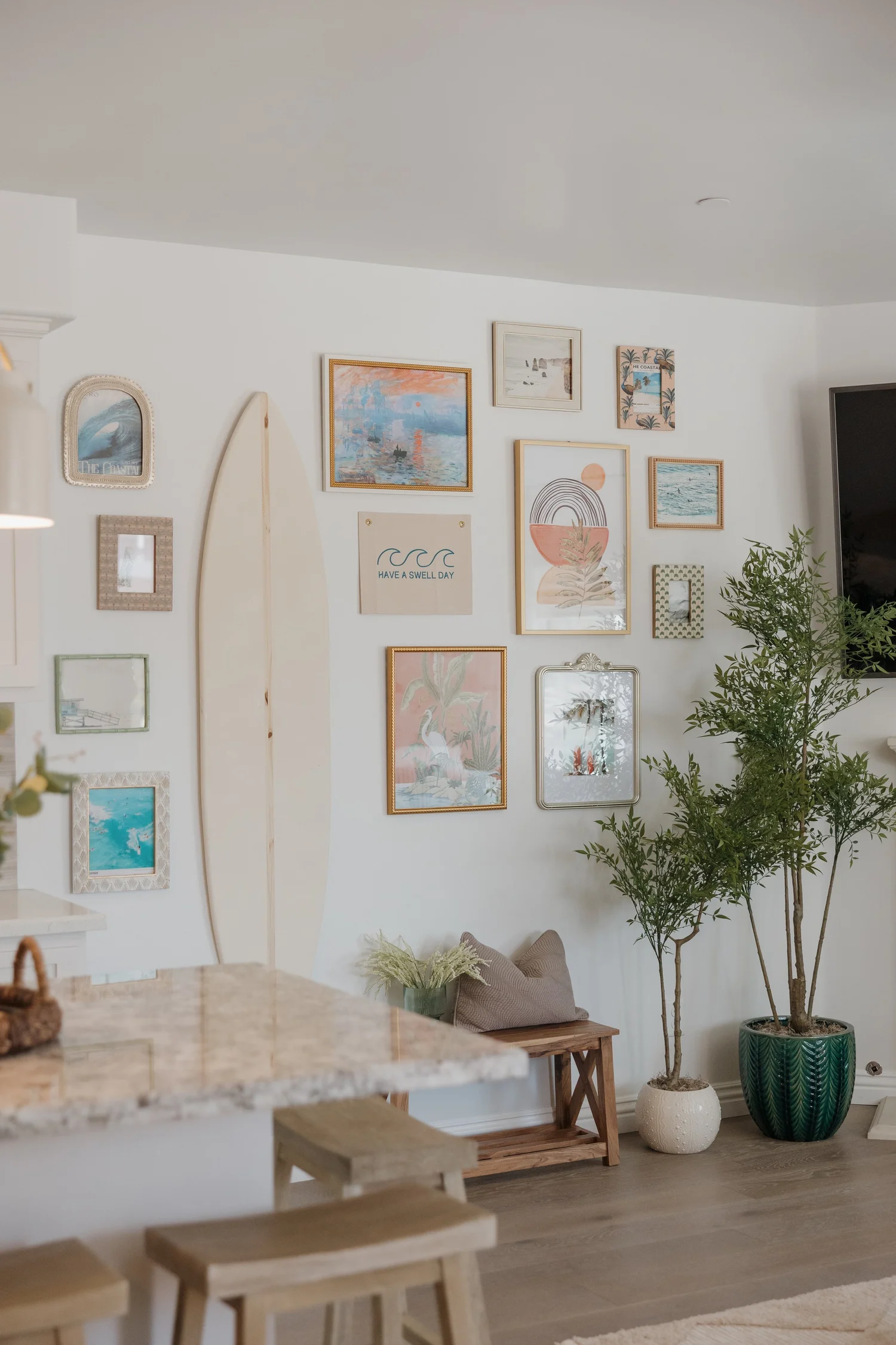 Gallery wall of coastal art and a surfboard beside the kitchen counter at Coastal Palm in Pacific Beach, designed by Salt and Sky Lodging Co.