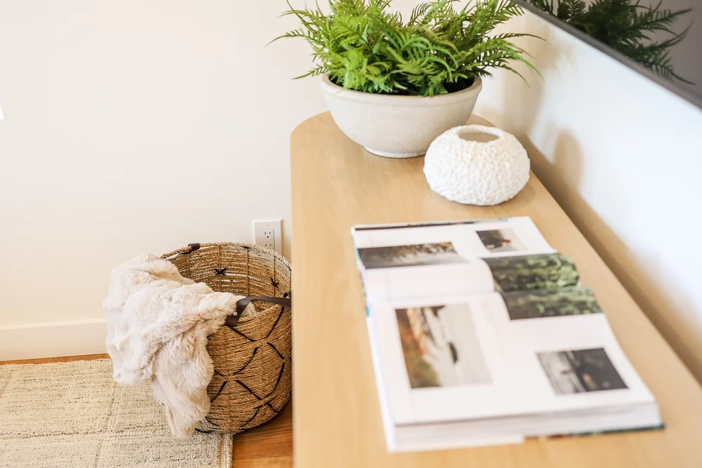 A coffee table book, a fern and a woven basket by the media console in Unit Three at The Swell, Oceanside