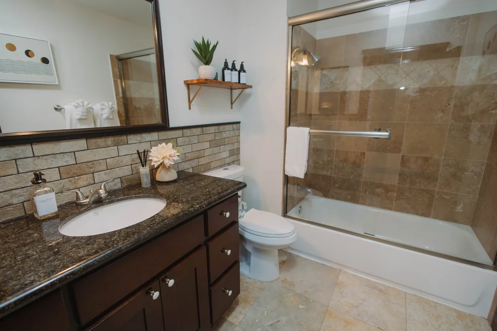 The upstairs hall bathroom at Palm Parkside, with a granite vanity, a brick backsplash and a tub with a glass shower door