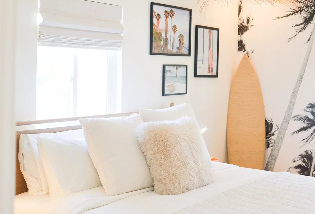 A bedroom at The Swell Unit Three in Oceanside, with a palm mural, a woven pendant and a surfboard in the corner