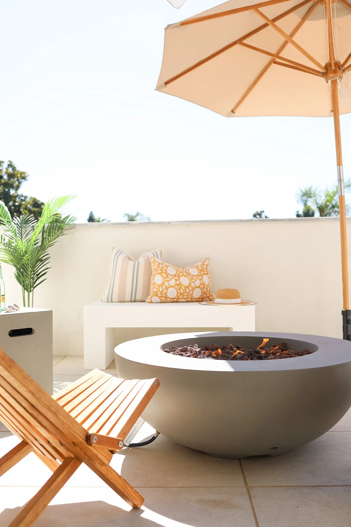 The sunny patio at The Swell, with a round fire table, a teak folding chair, a cushioned bench and a market umbrella