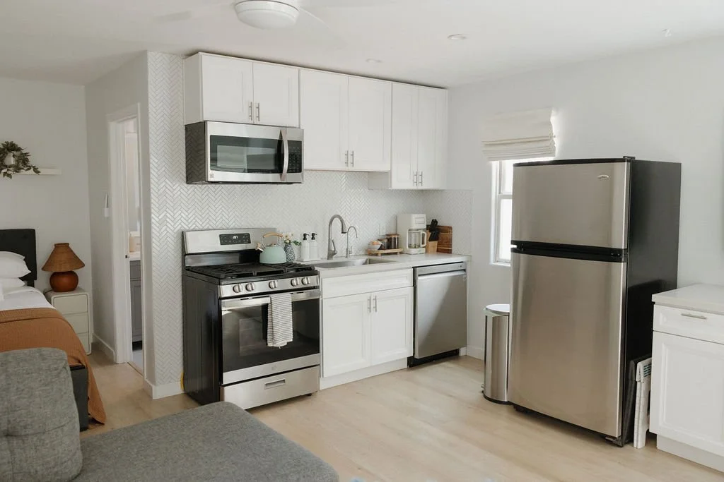 Small kitchen area with white cabinets, stainless steel appliances including a microwave, stove, dishwasher, and refrigerator. Light wood flooring, a small window with blinds, and minimal decor.