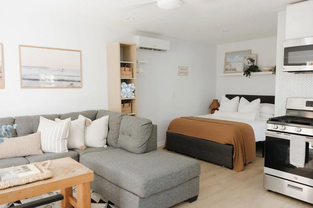 Cozy studio apartment with a gray sectional sofa, decorative pillows, a wooden coffee table, a bed with white linens and a brown throw, a small wooden nightstand with a lamp, a built-in open shelf, artwork on the walls, a microwave above a stove, and