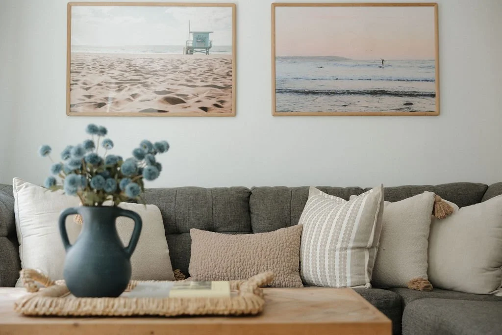 Living room with a gray sectional sofa decorated with white and beige pillows, wooden coffee table with a blue vase and flowers, and two beach-themed framed pictures on the white wall.