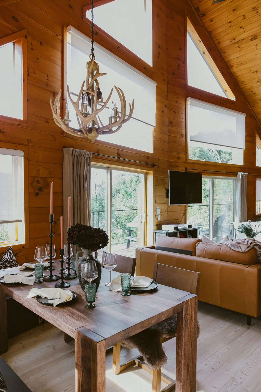 Interior of a cozy wooden cabin living room and dining area with large windows, a chandelier made of antlers, a dining table set with plates, glasses, and candles, and a leather sofa with a wall-mounted TV.