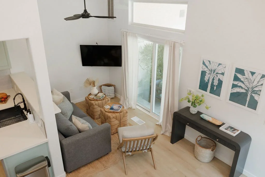 A living room with a gray sofa, a wall-mounted flat-screen TV, a large window with white curtains, a black console table with a vase of green flowers, and two framed botanical prints. There is a sliding glass door leading outside, and a ceiling fan v