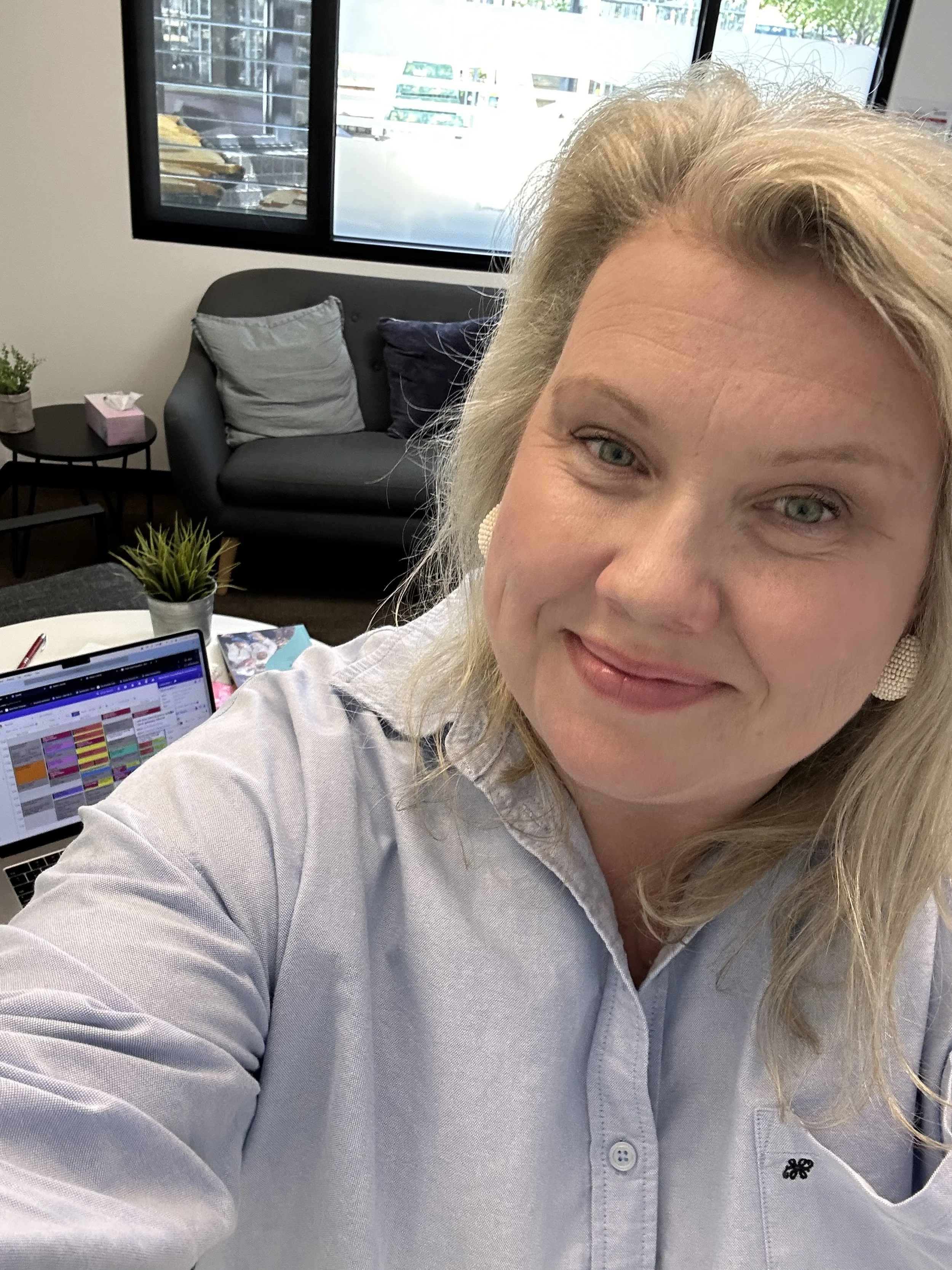 A woman with blonde hair taking a selfie in an office space. Behind her, there is a black couch with gray and black pillows, a small side table with a pink tissue box and a potted plant, and a window showing a parking lot and outdoor scenery. A laptop with colorful spreadsheet is open on the desk.