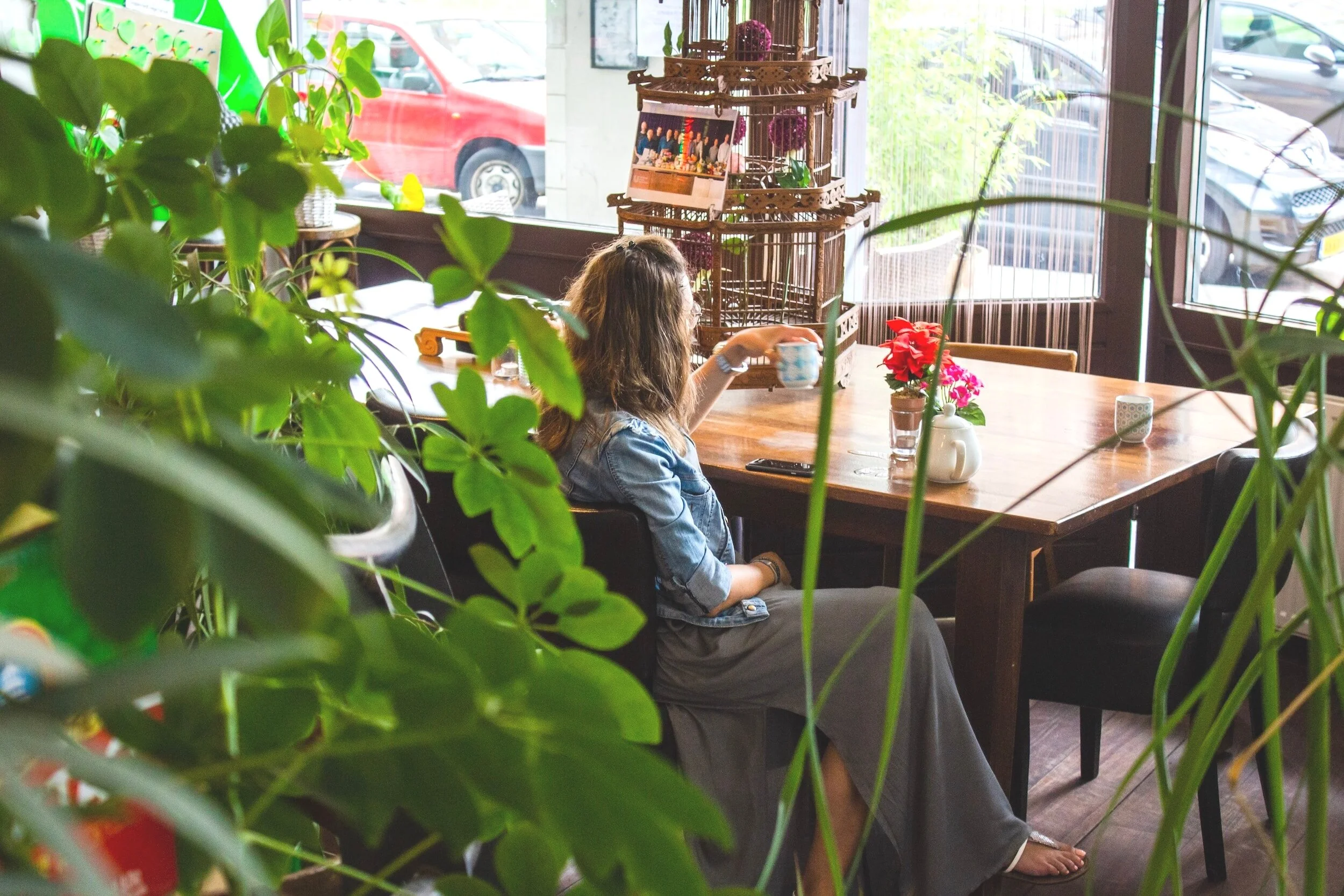 A woman sitting at a wooden table inside a cafe, surrounded by green plants.  The table has a teapot, a cup, and some flowers. Large windows show parked cars outside. She is alone and thinking.