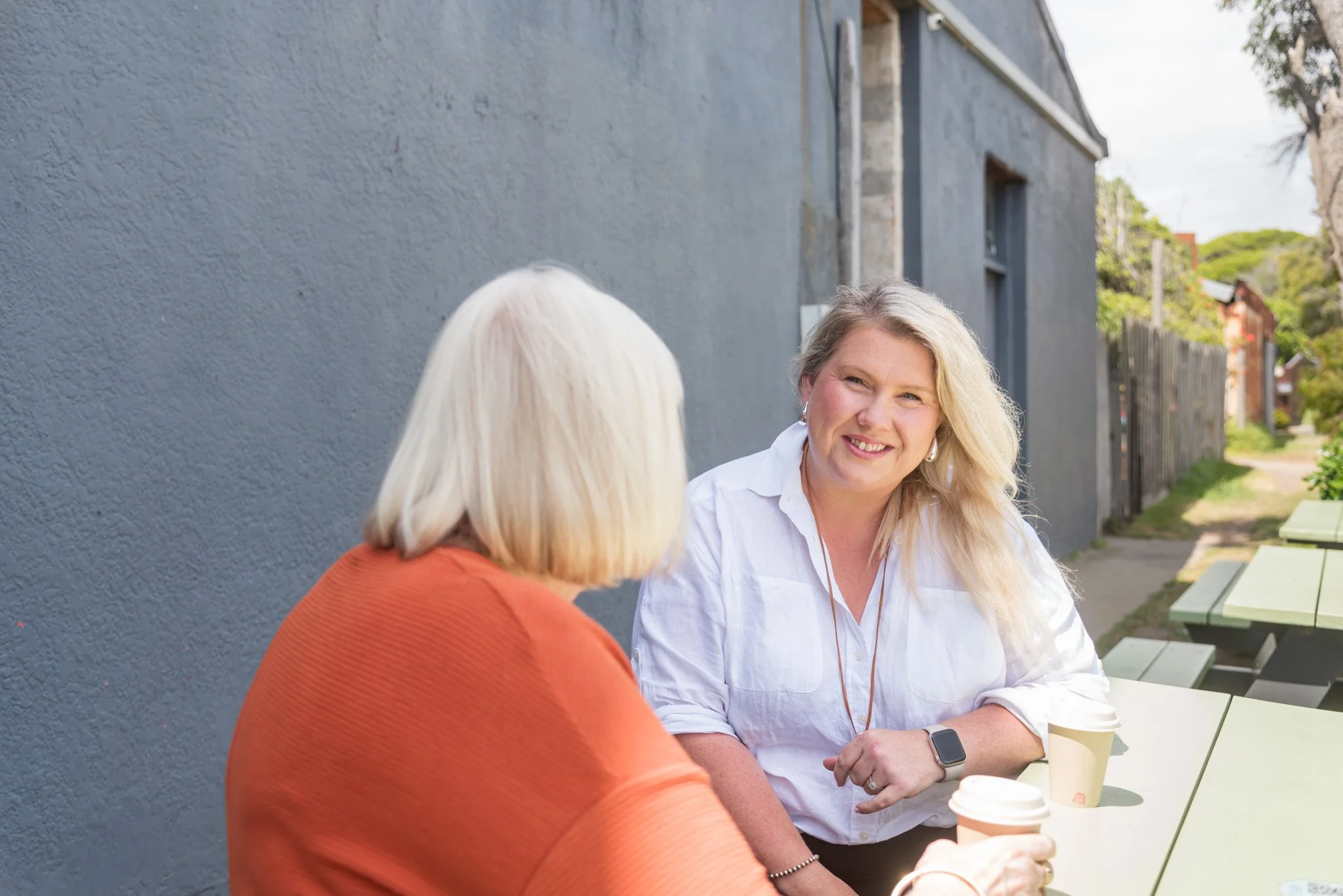 Two women are sitting at an outdoor table, engaged in a conversation, with coffee cups in front of them. The woman facing the camera has blonde hair and is smiling, while the other woman's back is visible, showing white hair and an orange sweater. They are sitting against a gray wall with trees and houses visible in the background.
