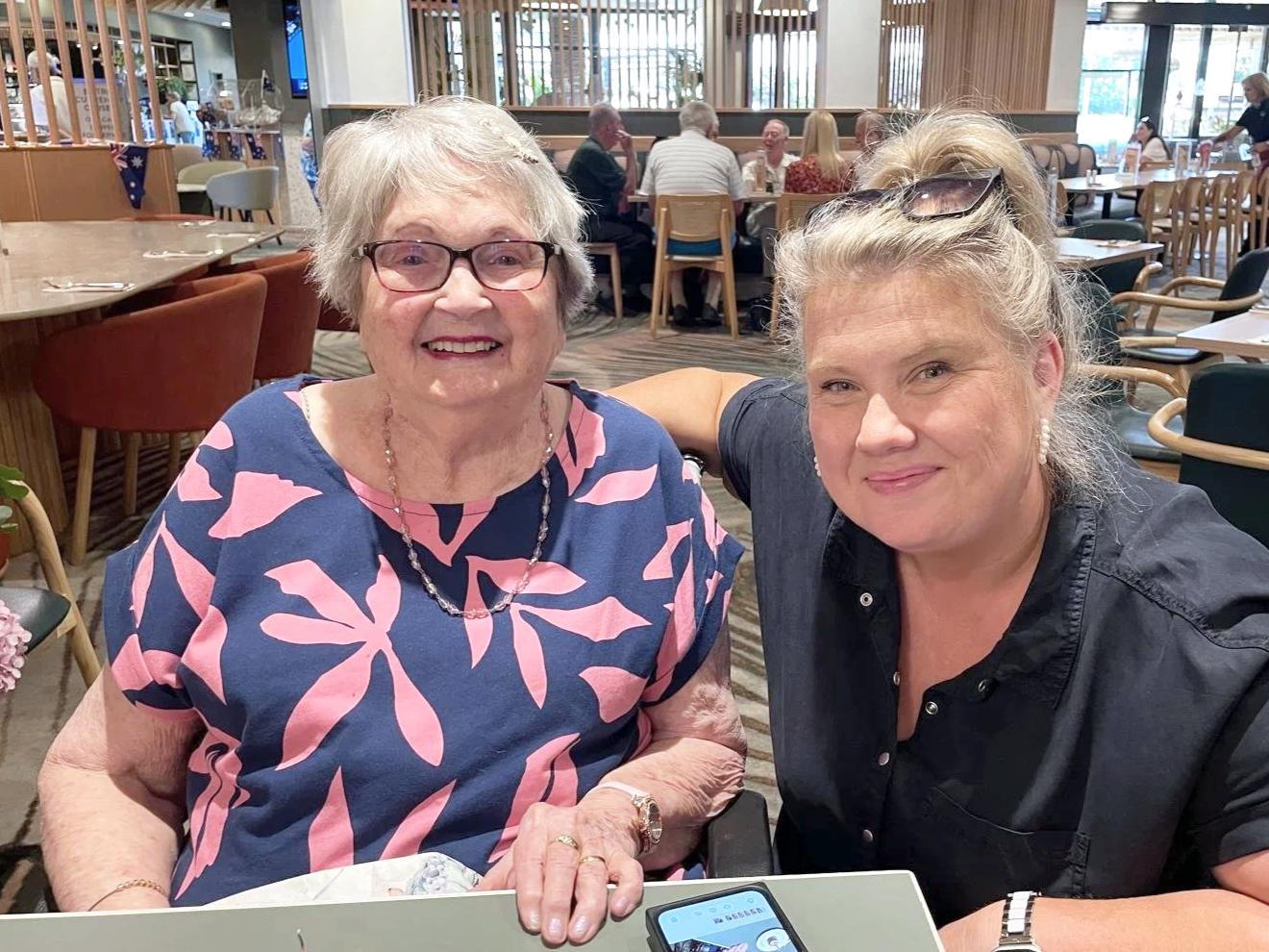 Two women smiling at a restaurant table, one elderly with glasses and a patterned dress, the other middle-aged in a black shirt, with other people in the background.