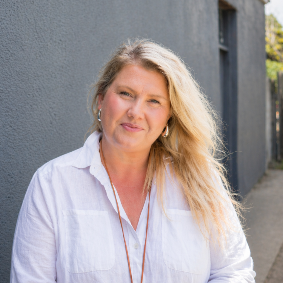 A woman with blonde hair, wearing a white shirt and hoop earrings, standing outdoors against a gray wall, smiling.