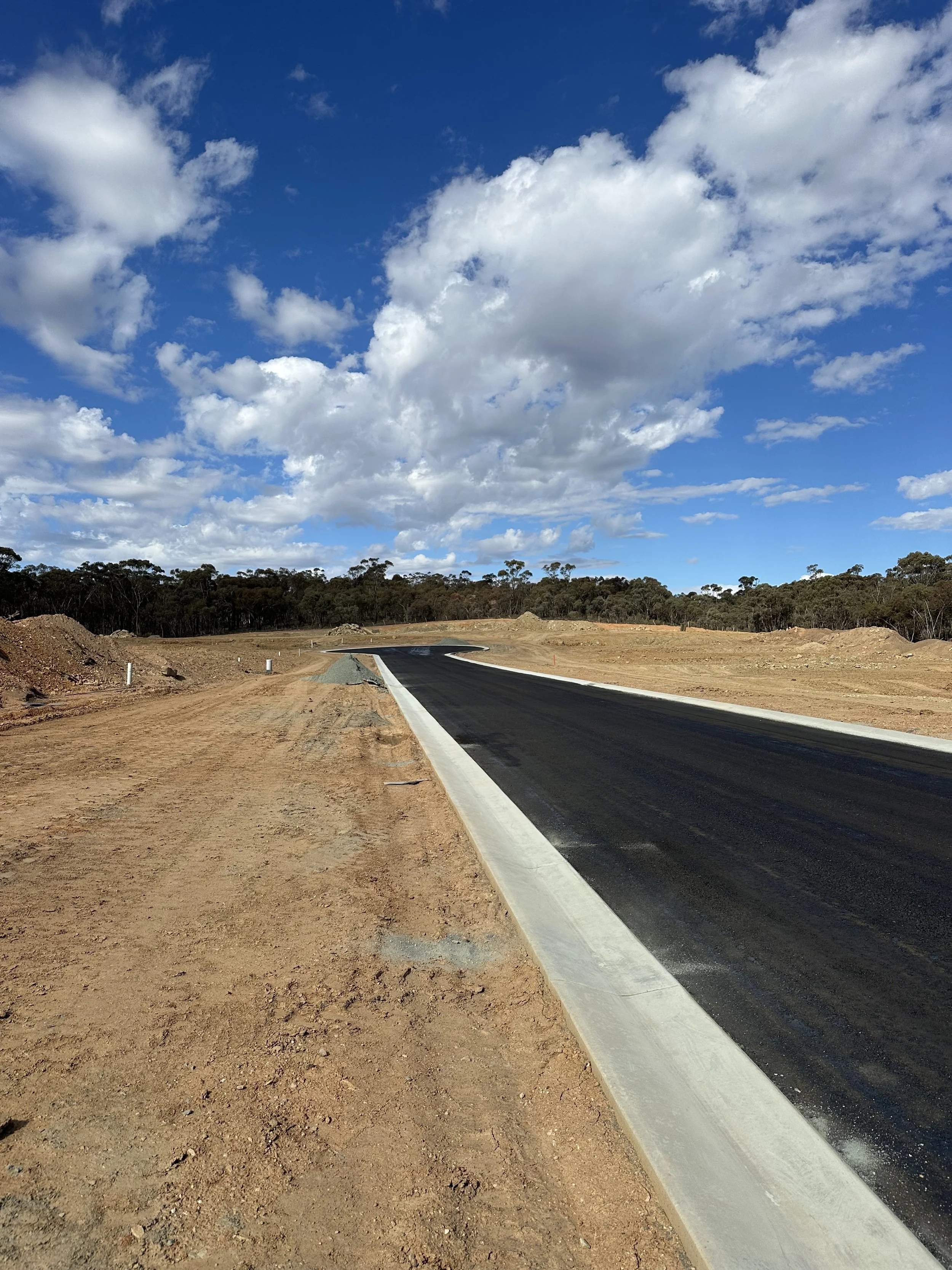 A newly paved asphalt road winding through a rural landscape with trees in the background, under a partly cloudy sky.