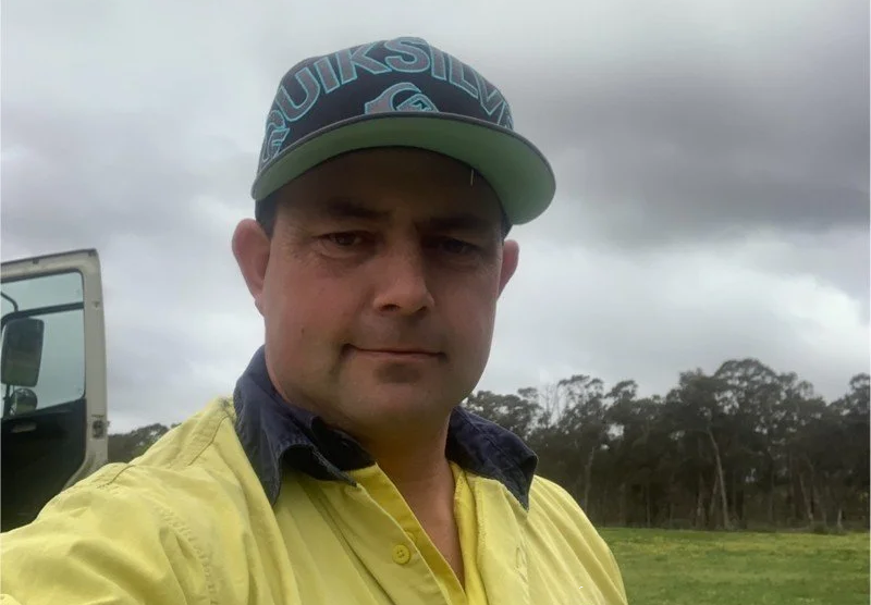 A man taking a selfie outdoors, wearing a baseball cap and a yellow shirt, with trees and a cloudy sky in the background.
