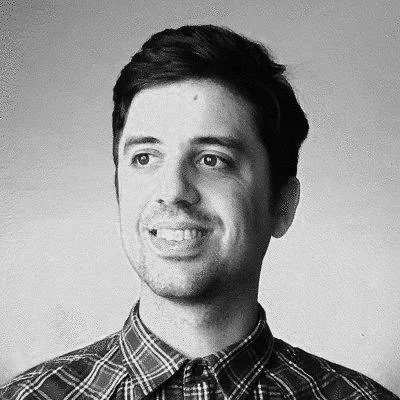 A young man with dark hair, wearing a checkered shirt, smiling and looking to the side, in black and white.