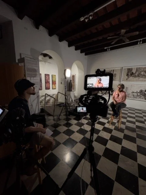 A video interview setup in an art gallery with a person being filmed and a crew member managing the camera.