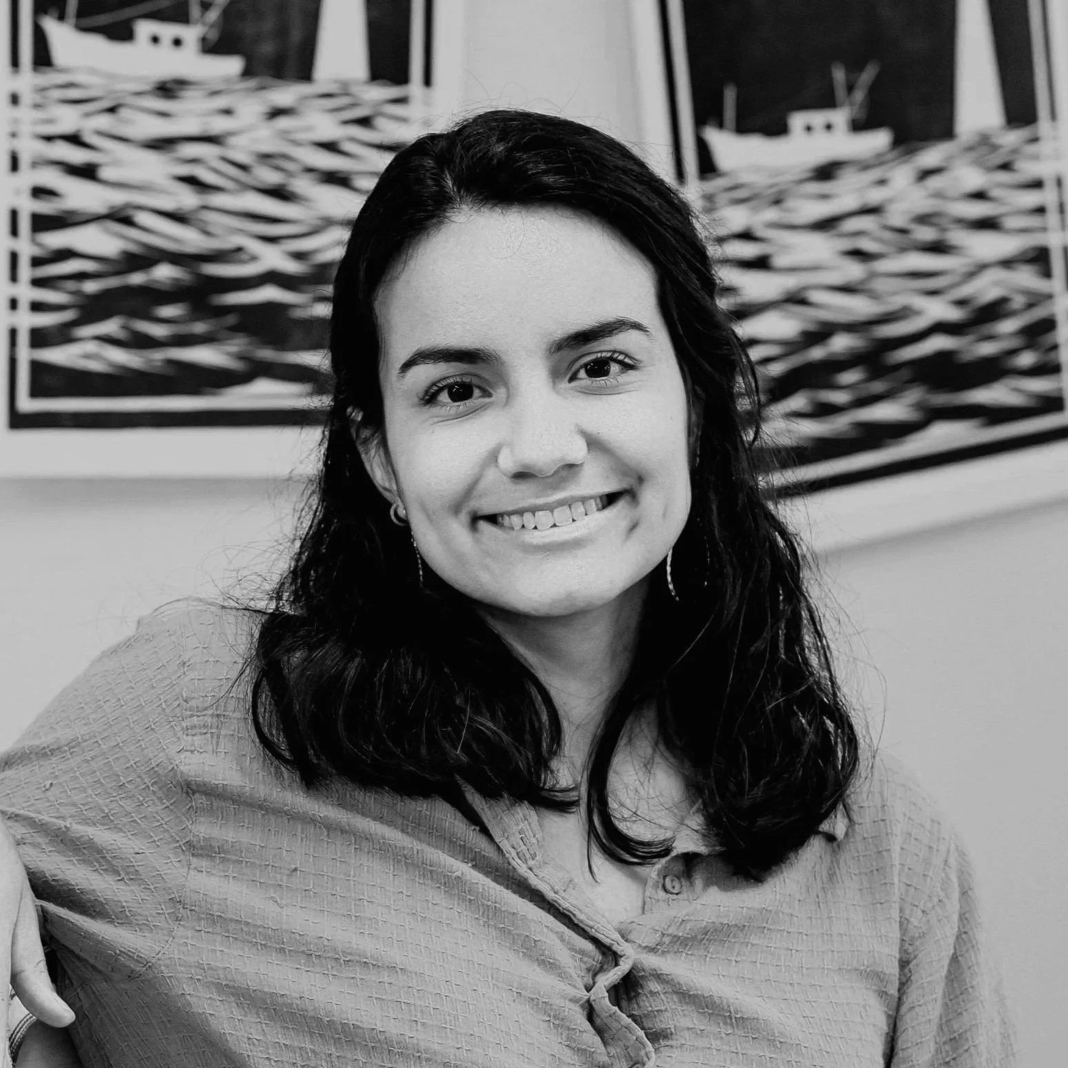 Black and white photo of a smiling woman with dark hair, wearing earrings, sitting in front of abstract nautical paintings.
