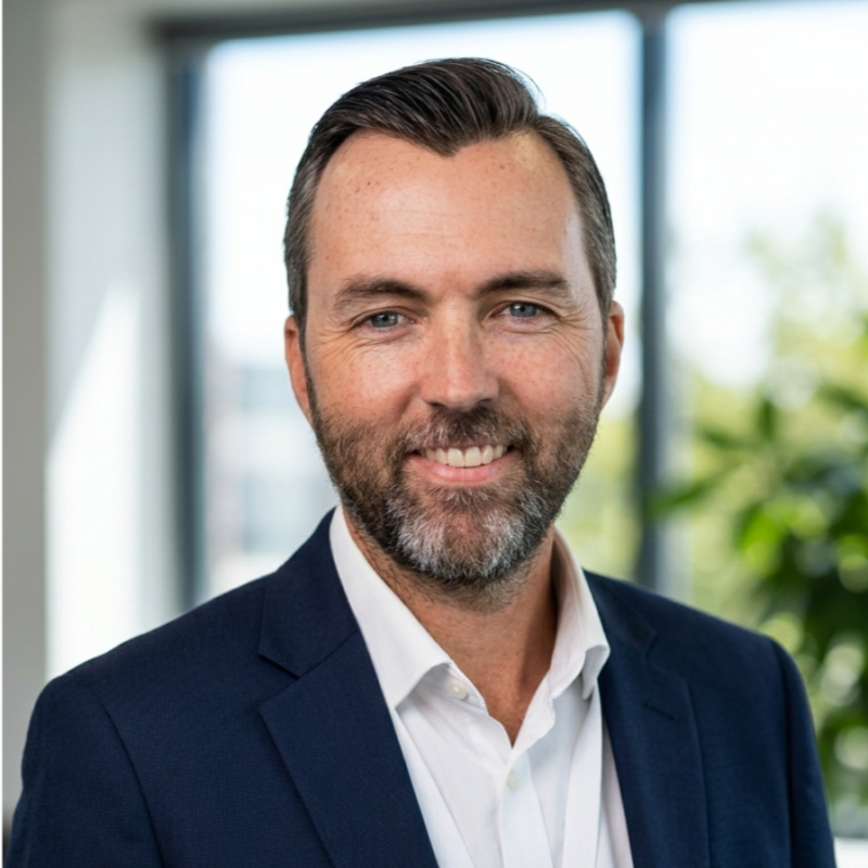 Professional portrait of a middle-aged man with short brown hair and a beard, wearing a navy blazer and a white shirt, in an office setting with large windows and greenery outside.