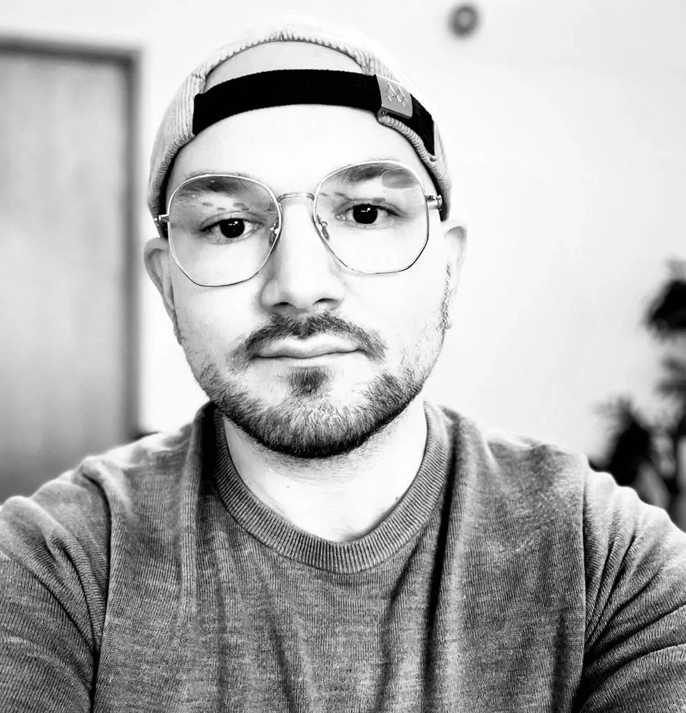 A young man with a beard and mustache wearing a baseball cap backwards, large round glasses, and a casual t-shirt, sitting indoors, with a blurred background.