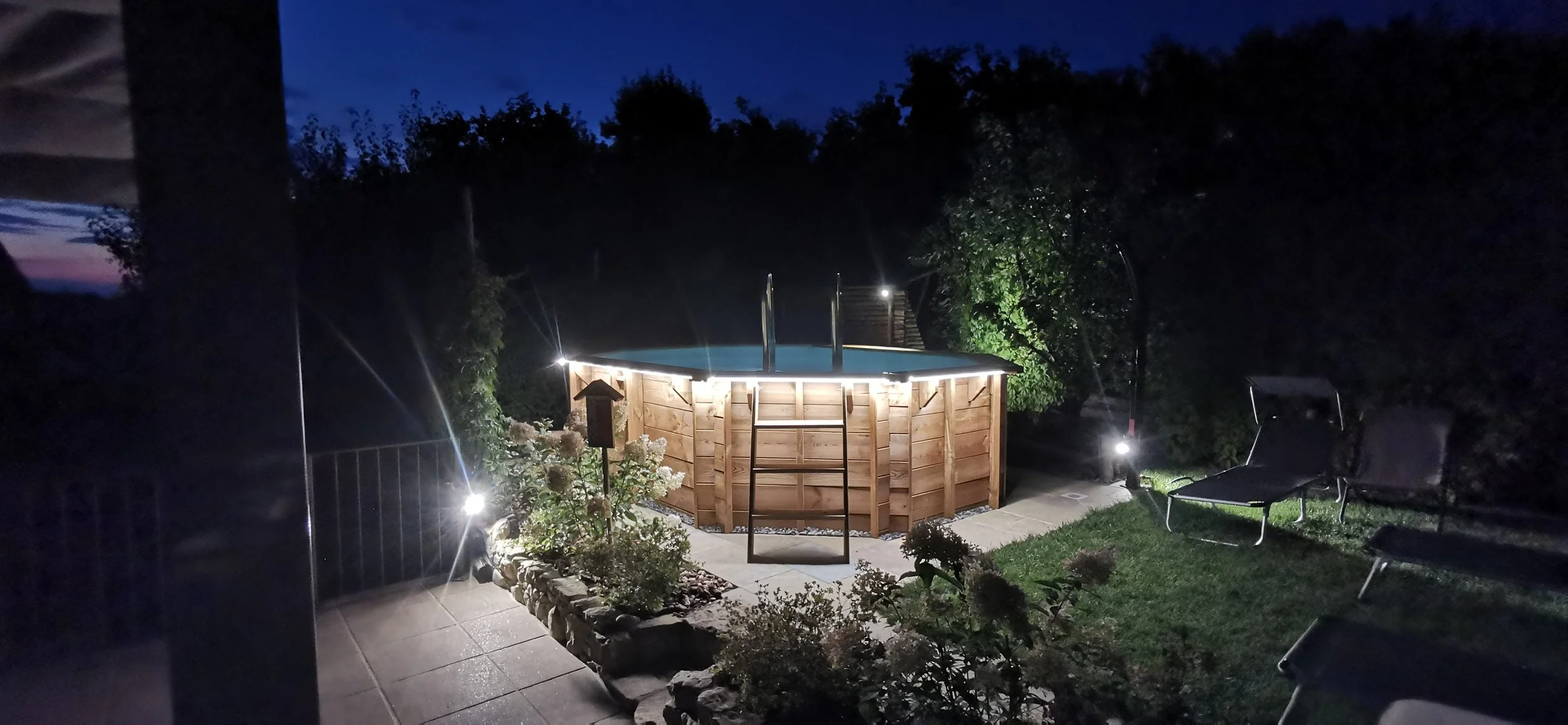 Piscina gonfiabile in legno con scaletta, illuminata di notte, in un giardino con panchine e piante, sotto un cielo al tramonto o crepuscolo.