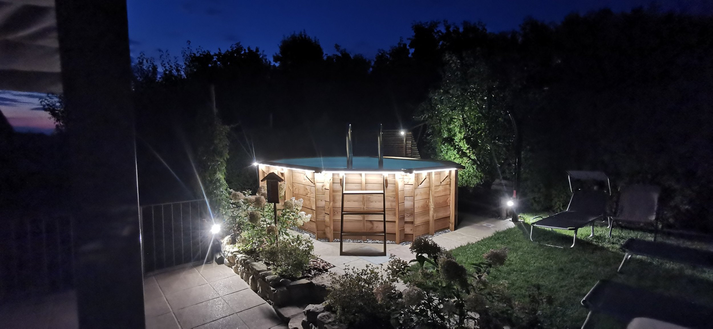 Piscina fuori terra in legno illuminata di notte, circondata da sedie a sdraio e piante, con un cielo scuro sullo sfondo.