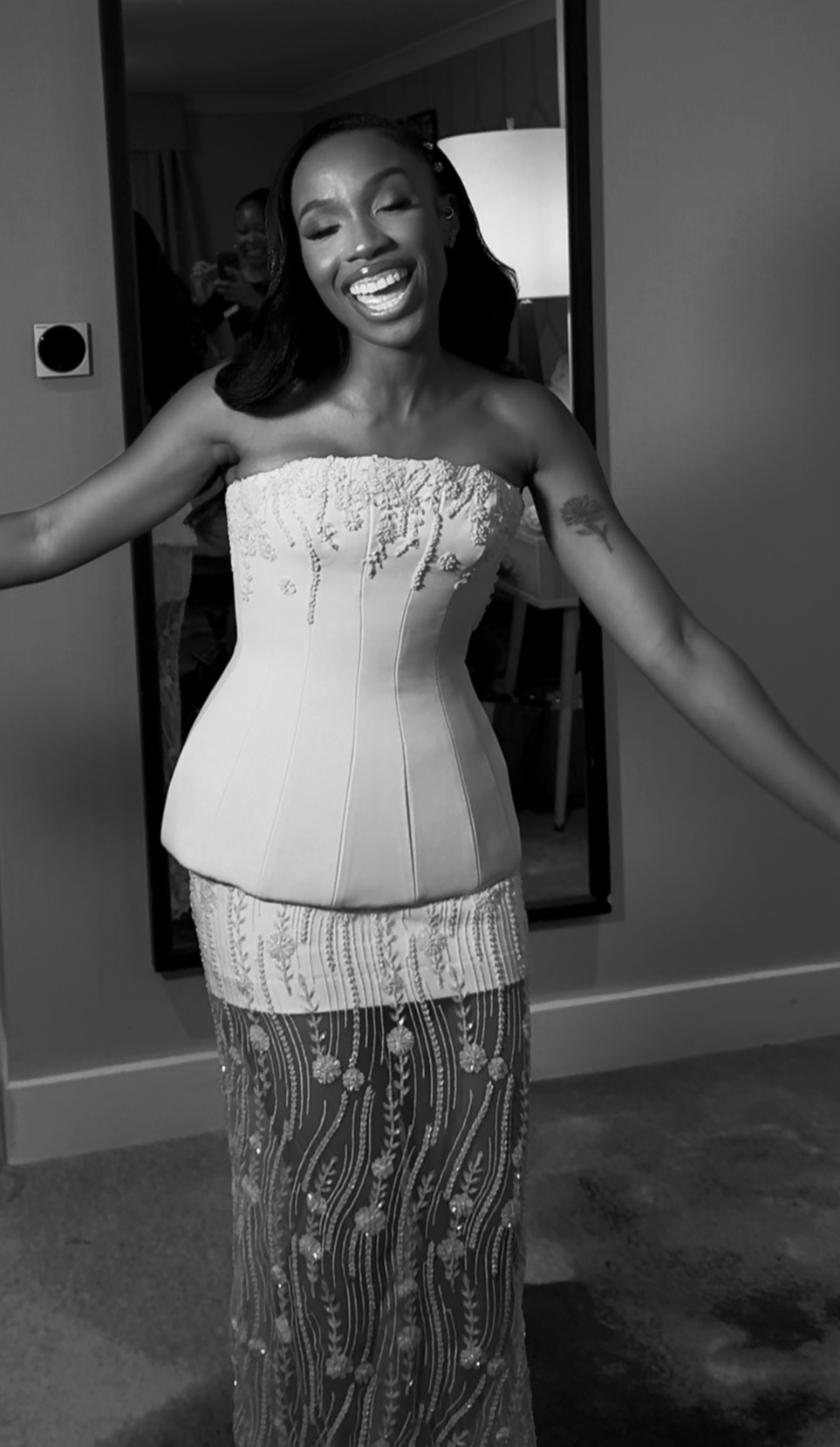 A woman in a strapless dress smiling with her arms outstretched, standing in a room with a mirror behind her reflecting other people.