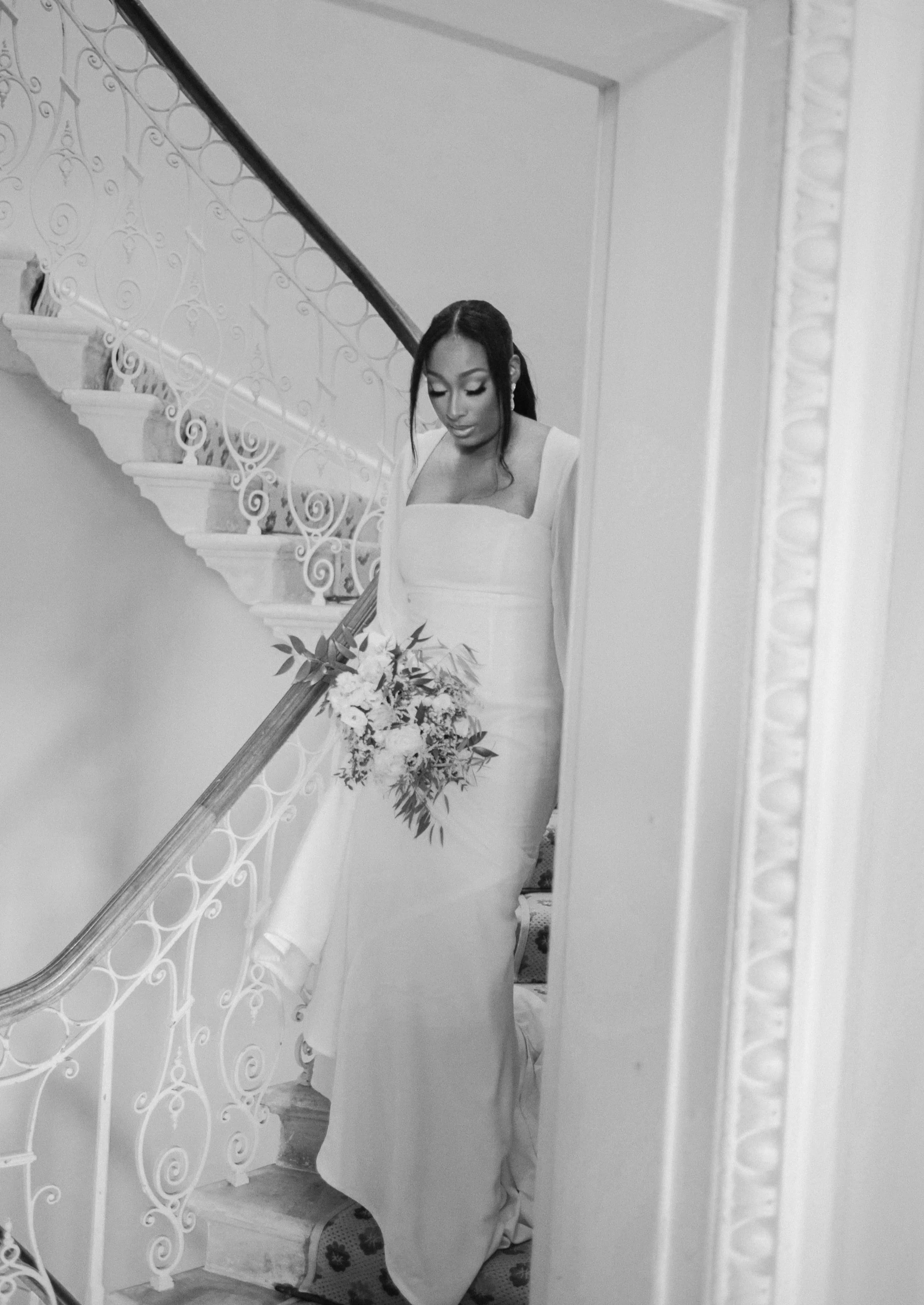 A bride in a white wedding dress holding a bouquet of flowers, standing on stairs inside a house, looking down.