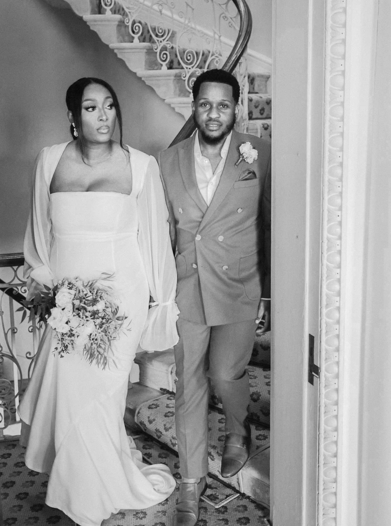 Black-and-white photo of a bride and groom standing side by side in an elegant interior, with a staircase in the background. The bride is holding a bouquet and wearing a wedding dress with long sleeves, while the groom is dressed in a double-breasted