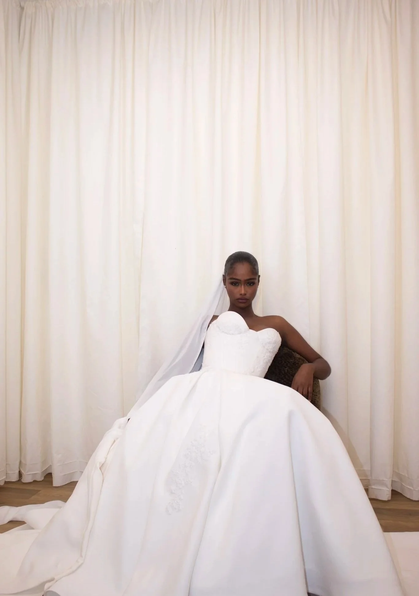 A woman in a wedding dress sitting in front of light-colored curtains.