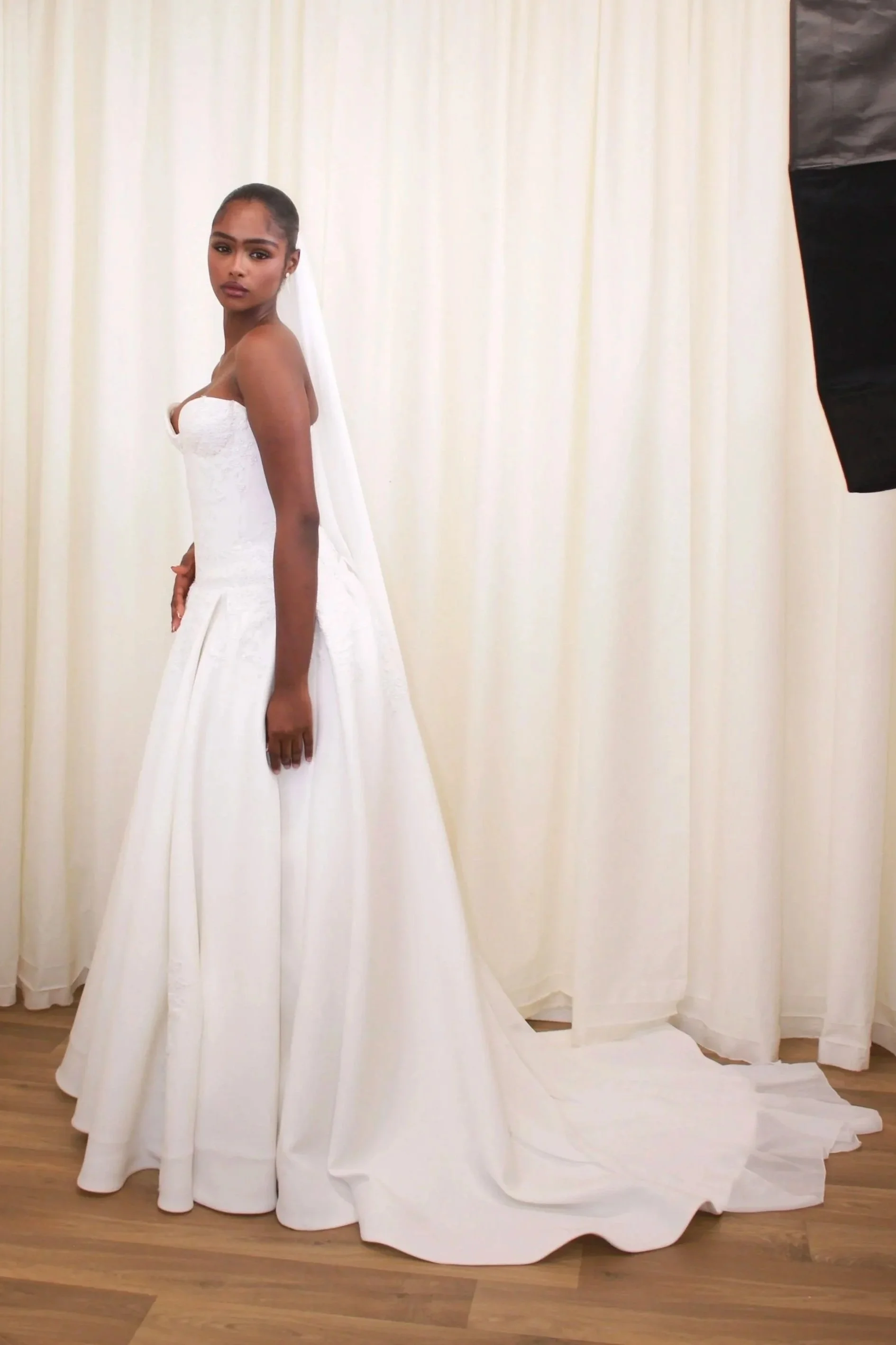 A woman in a white strapless wedding dress with a long train, standing in front of cream-colored curtains.